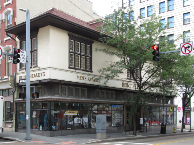 Eine belebte Stadtstraße mit einem geparkten Bus, Gebäuden, Bäumen, Laternen, Verkehrsampeln, Werbetafeln und einem Kaufhaus mit Schaufensterpuppen im Hintergrund unter einem sichtbaren Himmel.