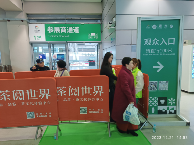 Eine Gruppe von Menschen steht vor einem grünen Teppich auf einer Tee-Messe in China, einige halten Taschen, Schilder und Plakate mit Text, eine Flasche auf dem Boden, ein Display, Metallstangen, eine Überwachungskamera an der Wand und eine Decke mit Deckenleuchten.