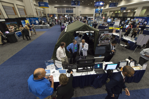 Eine große Gruppe von Menschen auf einer Karriere-Messe um einen Tisch mit Laptops, Monitoren und Papieren versammelt, mit einem Zelt, Schildern, Deckenleuchten und Fenstern im Hintergrund.