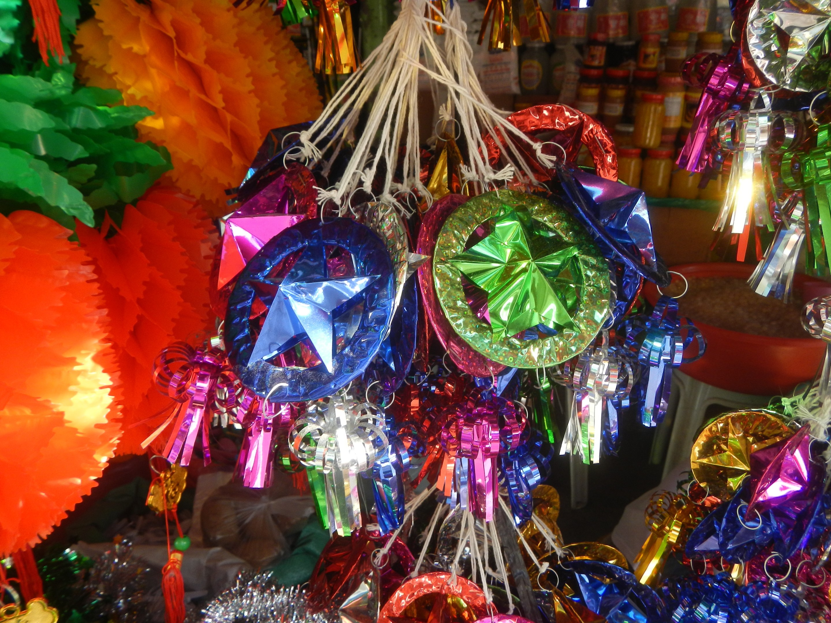 Farbenfrohe Festschmuck-Dekorationen, darunter Laternen und Bänder, hängen von der Decke über Regalen mit Flaschen im Hintergrund.
