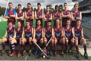Eine Gruppe von Männern in Sportbekleidung steht auf einem Feld und hält Hockey-Schläger, feiernd mit einem Stadion und Zuschauern im Hintergrund.