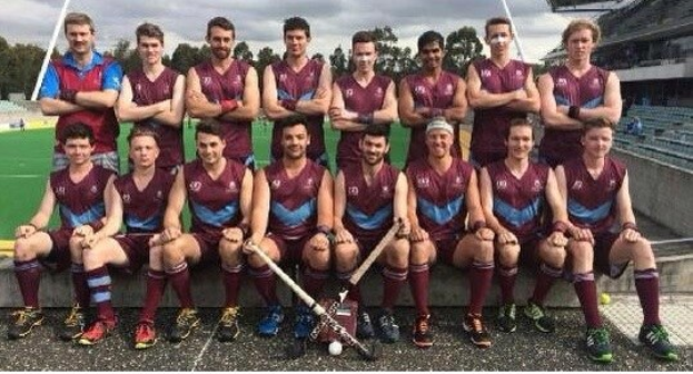 Eine Gruppe von Männern in Sportbekleidung steht auf einem Feld und hält Hockey-Schläger, feiernd mit einem Stadion und Zuschauern im Hintergrund.