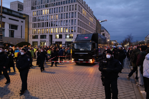Eine Gruppe von Menschen steht in der Nähe eines Lastwagens auf einer Straße, die von Gebäuden, Bäumen und Laternen gesäumt ist, unter einem bewölkten Himmel, wobei einige eine Mütze und eine Maske tragen und ein Band an einem Pfahl im Vordergrund.