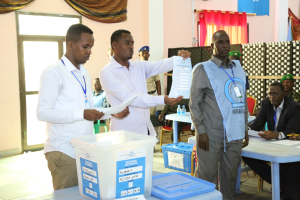Eine Gruppe von Männern steht um einen Tisch mit verschiedenen Gegenständen herum und stimmt bei einer Wahlurne ab, wobei im Hintergrund Personen und Möbel zu sehen sind.
