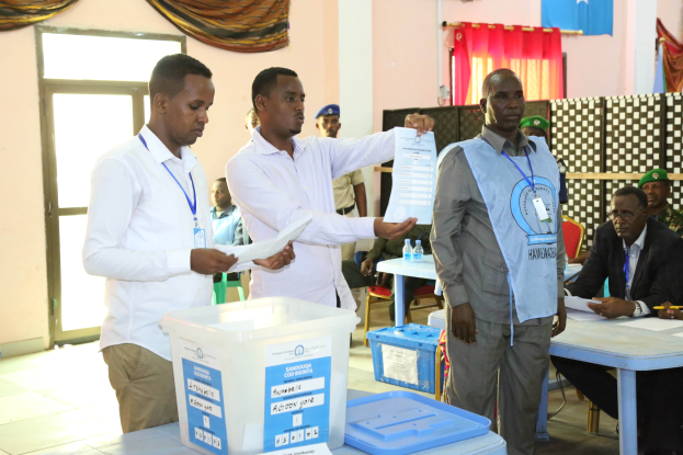 Eine Gruppe von Männern steht um einen Tisch mit verschiedenen Gegenständen herum und stimmt bei einer Wahlurne ab, wobei im Hintergrund Personen und Möbel zu sehen sind.