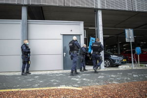 Eine Gruppe von Polizeibeamten steht in einer Parkplatz vor einem Gebäude mit einer Tür, umgeben von Fahrzeugen, Pfählen, Brettern und verstreuten Steinen.