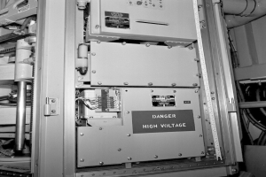 Ein Schwarz-Weiß-Foto eines elektrischen Schaltkastens mit einer Gefahrenzeichen, umgeben von verschiedenen Maschinen und Rohren.