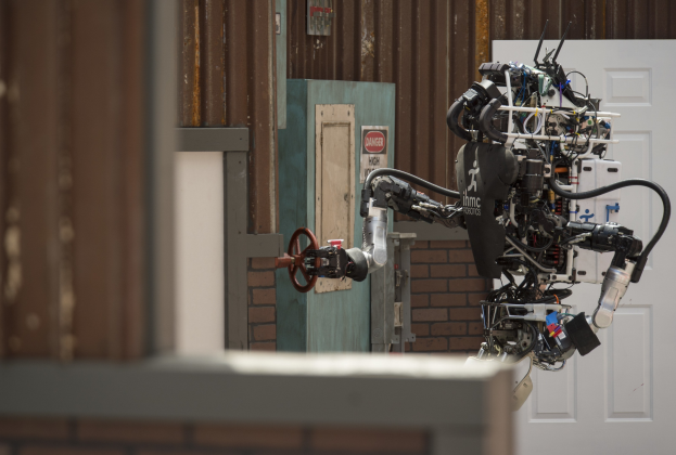 Ein humanoider Roboter mit ausgestreckten Armen und geneigtem Kopf steht vor einem Gebäude mit einer Tür und einer Wand im Hintergrund.